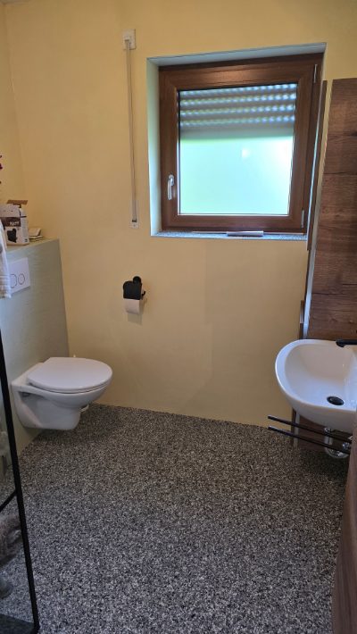Badezimmer mit grauem Steinteppich-Boden, Wand-WC und Waschtisch am Fenster – Badsanierung von mf.steintechnik aus Kirchheim am Neckar